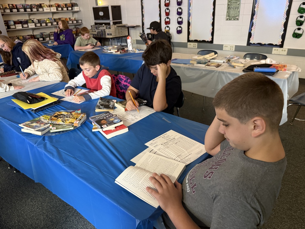 Students seated at blue table-covered desks in a classroom, quietly reading books and filling out worksheets.