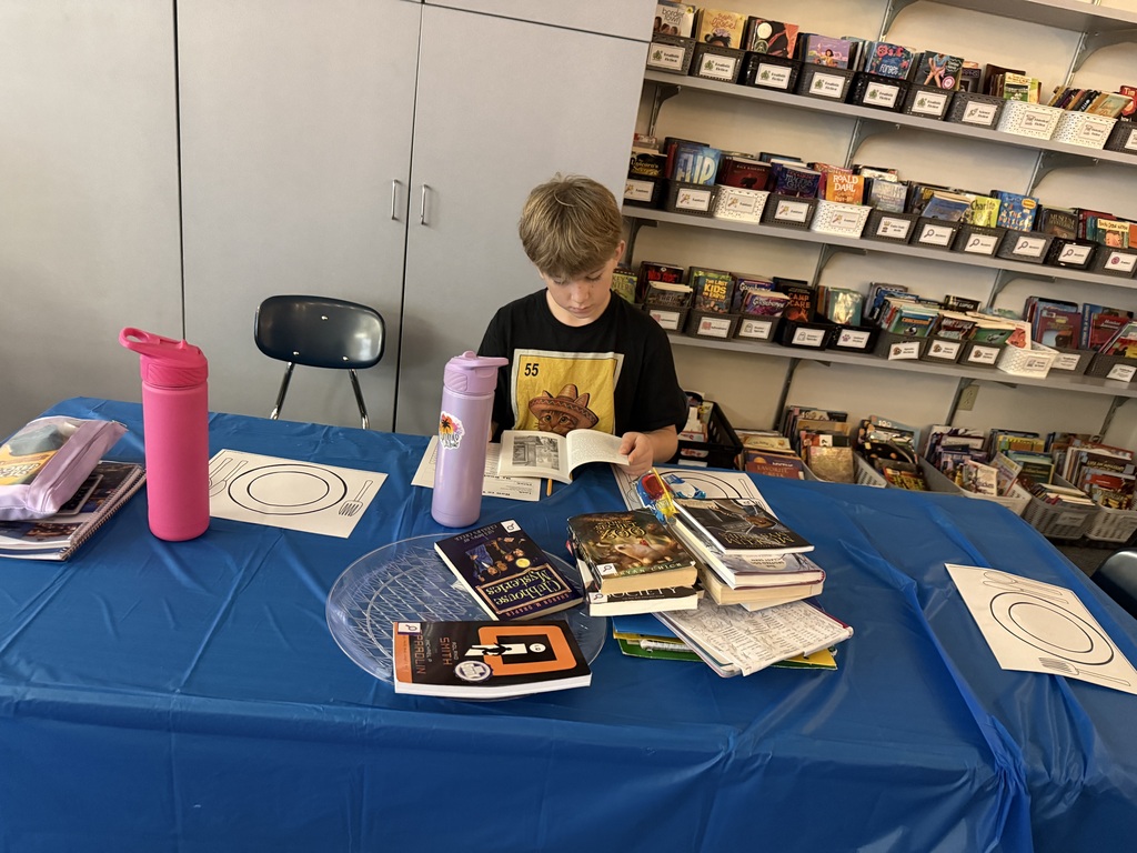 A young boy sitting alone at a blue table in a library classroom, focused on reading a book from a stack in front of him.