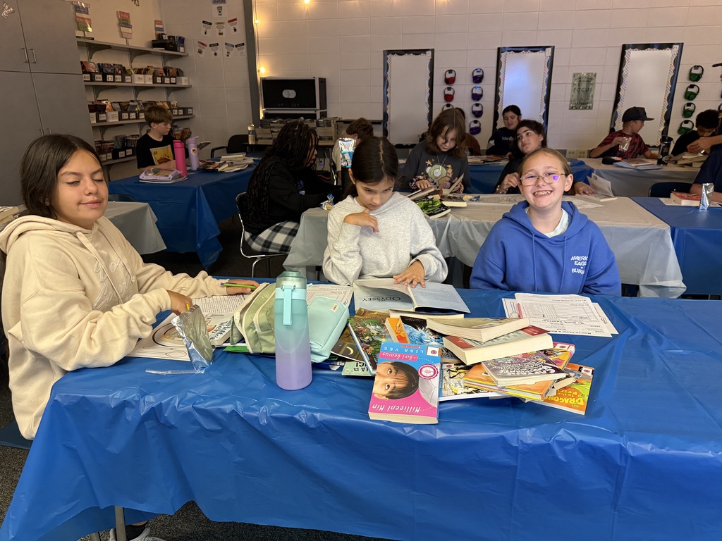 Three students sitting together at a blue table filled with books and school supplies, smiling and working during class.