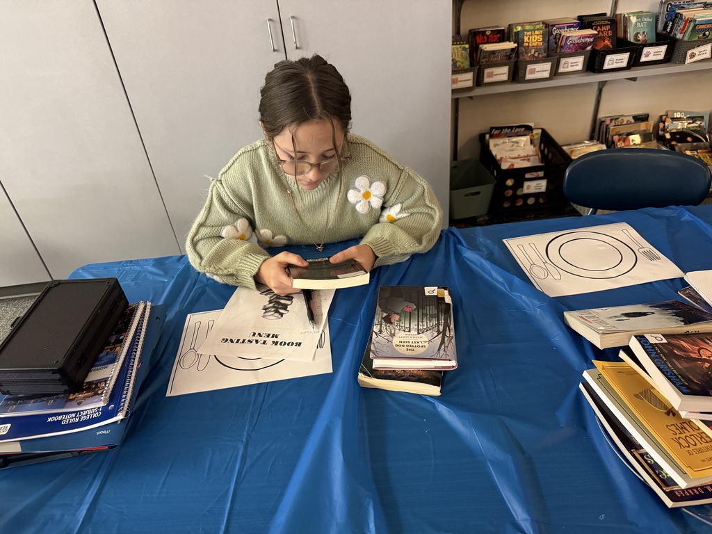 A student in a green sweater seated at a table covered in a blue tablecloth, attentively reading a paperback book.