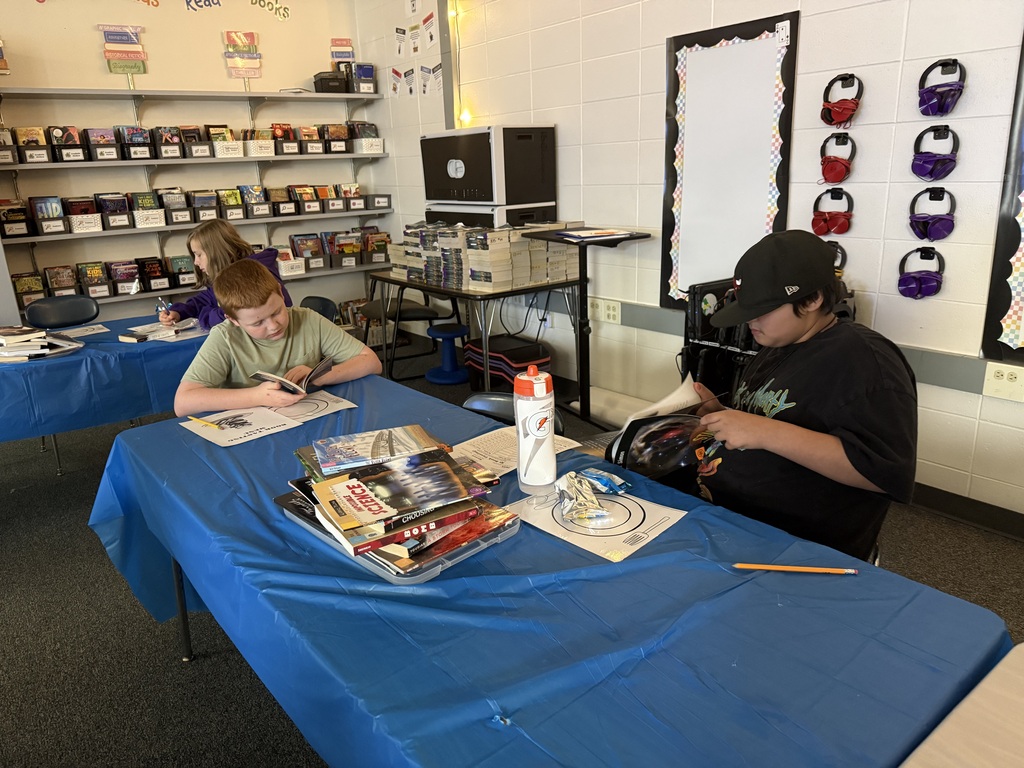 Two students sitting at a blue table in a classroom reading books, surrounded by stacks of paperbacks and school supplie