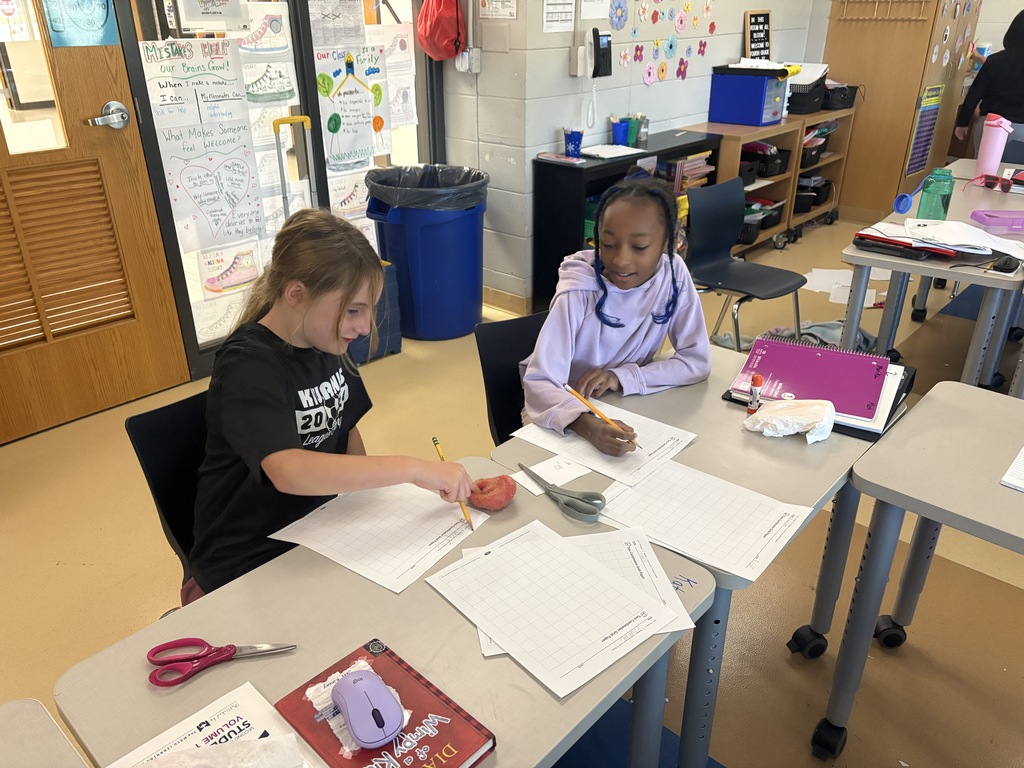 Two young female students working together at a classroom desk with worksheets, pencils, and school supplies.
