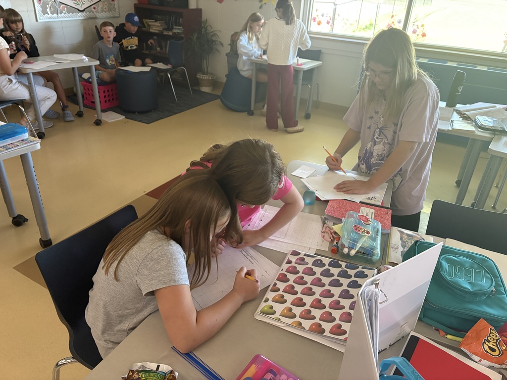 Elementary students collaborating and writing on worksheets at group tables inside a bright classroom.