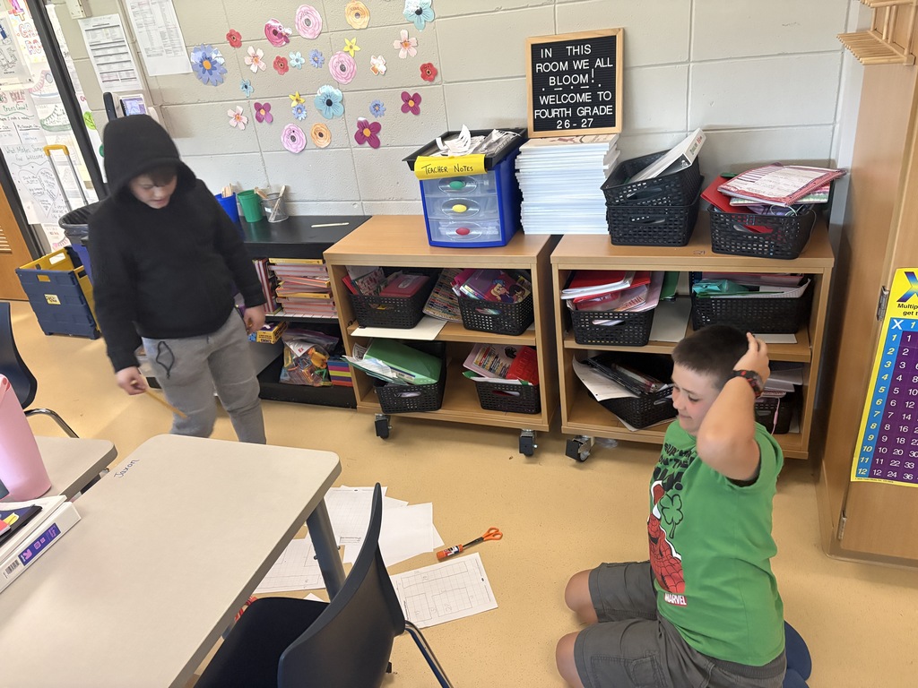 Two male elementary students participating in a classroom activity near storage shelves decorated with a "Welcome to Fourth Grade" sign.