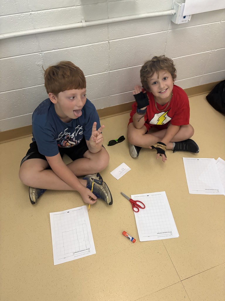 Two smiling young boys sitting cross-legged on the classroom floor working on paper assignments.