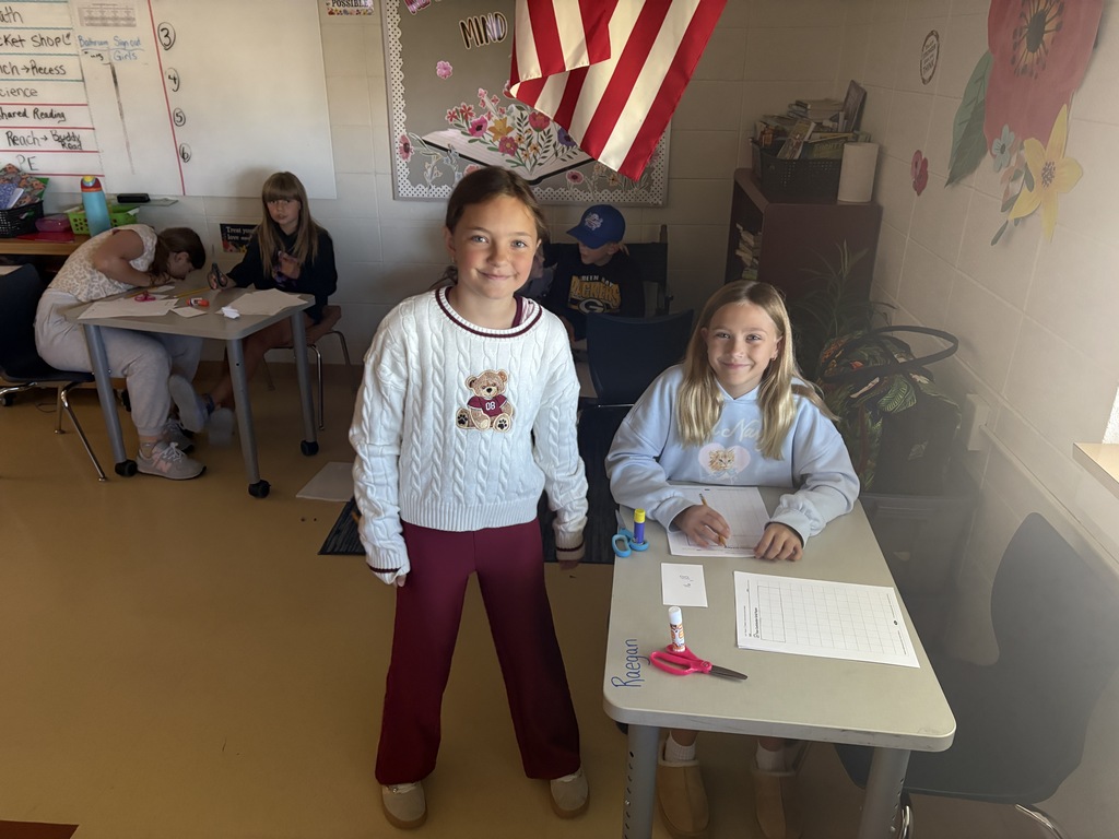 Two smiling female students at a classroom desk with schoolwork, with an American flag visible in the background.