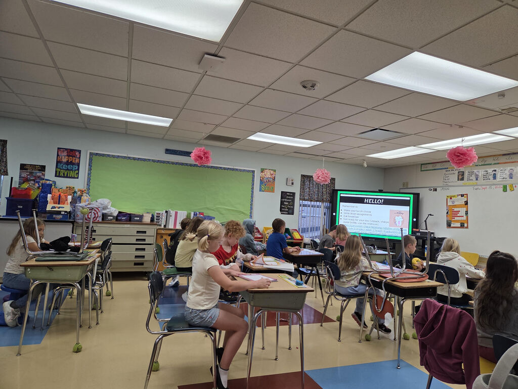 Students sitting at desks in a brightly lit classroom watching a presentation displayed on a smart screen at the front of the room.