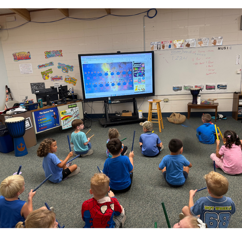 Elementary students sitting on a classroom floor, holding rhythm sticks while watching an interactive music lesson on a smart display board.