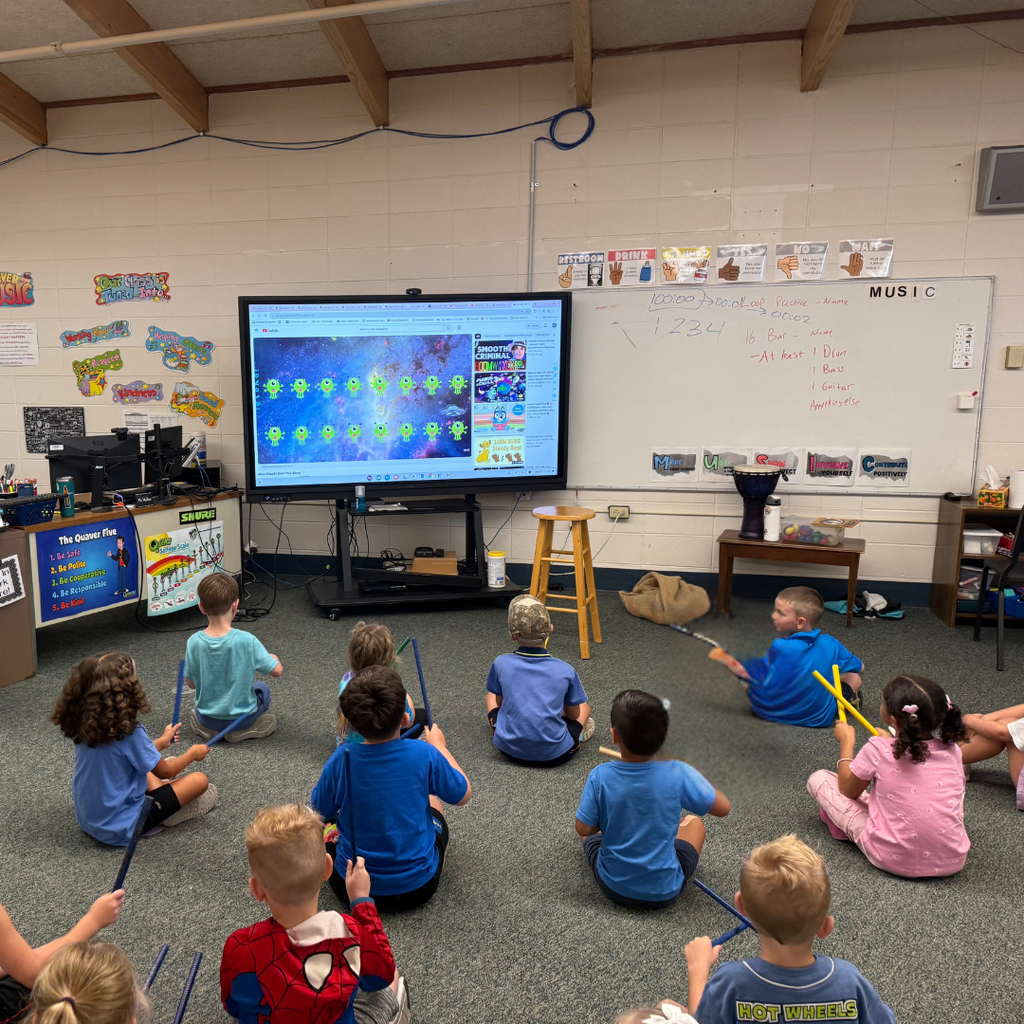 Young students seated on a carpeted music classroom floor, participating in a lesson with drumsticks in front of a digital screen.