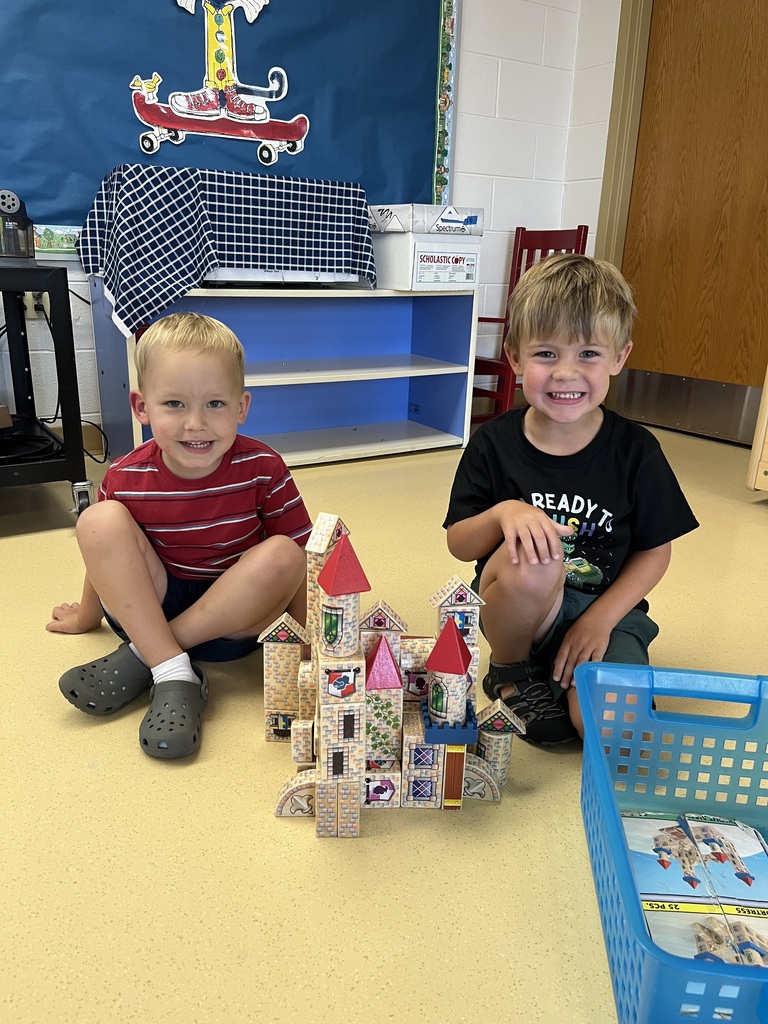 Two smiling young boys sit on a classroom floor next to a toy castle they built out of wooden blocks.