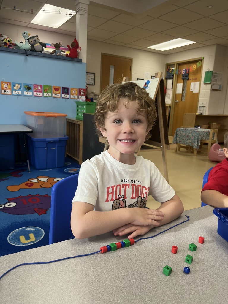A young student smiles brightly at a classroom table while stringing colorful beads on a blue cord.