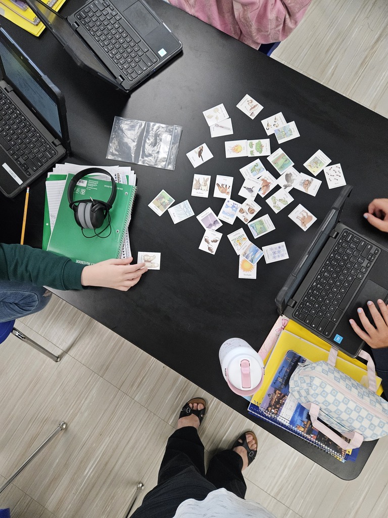 Overhead view of students collaborating at a dark classroom desk with laptops, notebooks, headphones, and science card-sorting picture cards spread out.