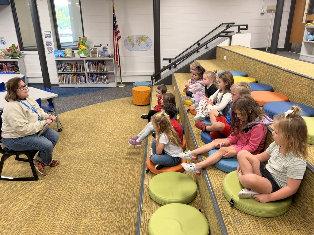 A teacher sits in front of young students sitting on colorful floor cushions on tiered seating in a school library listening attentively.