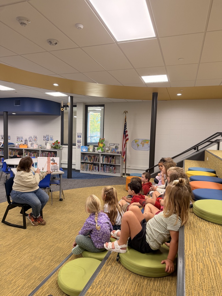 A teacher reads a picture book aloud to a group of young children sitting on round cushions on step seating inside a modern library