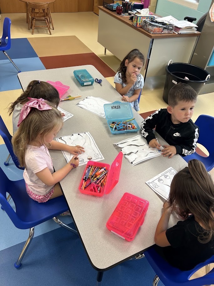 Group of young students sitting at a classroom table practicing fine motor skills with safety scissors, paper worksheets, and crayon boxes.