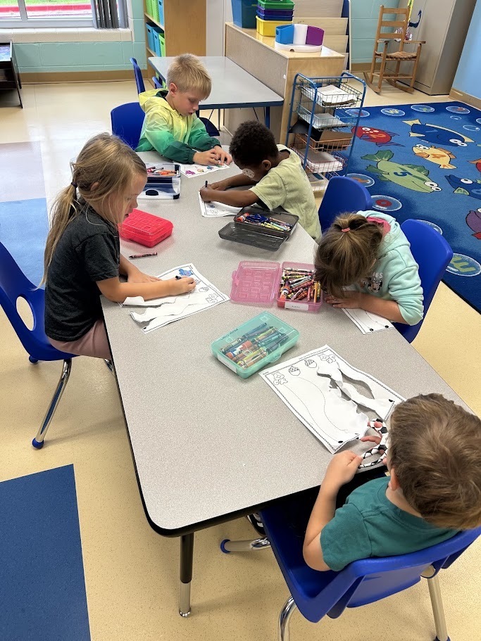 Young elementary students sitting around a classroom table working on paper cutting and coloring activities with crayons and pencil boxes