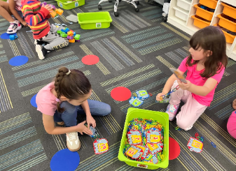 Two young students sitting on a classroom carpet sorting and connecting numbered activity cards next to a green storage bin.