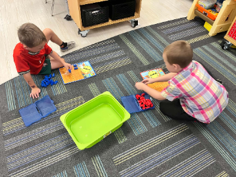 Two young students sitting on a classroom carpet using colorful game boards and plastic counting pieces next to a storage bin.