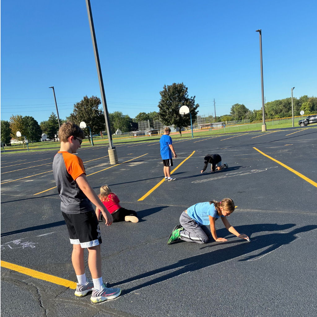 Elementary students working in pairs on a sunny school blacktop, tracing their shadows and drawing with sidewalk chalk.