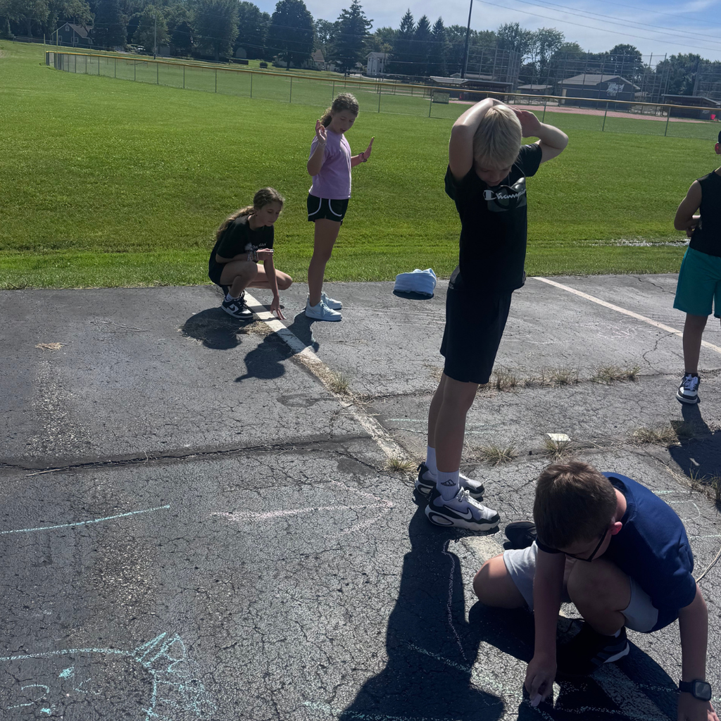 Elementary student standing with arms outstretched on a sunny blacktop as a classmate uses chalk to trace his shadow.