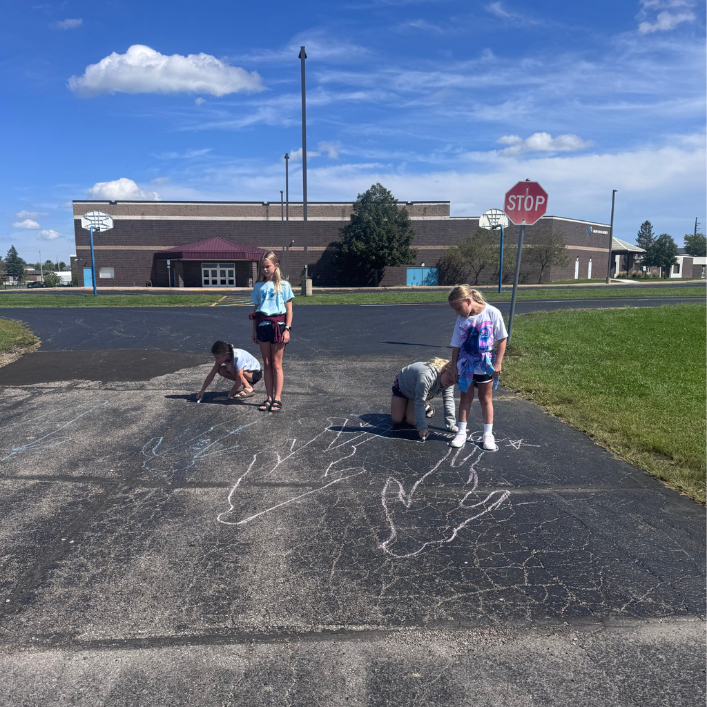 Students outside on a school blacktop tracing shadow outlines on the pavement with sidewalk chalk under a bright blue sky