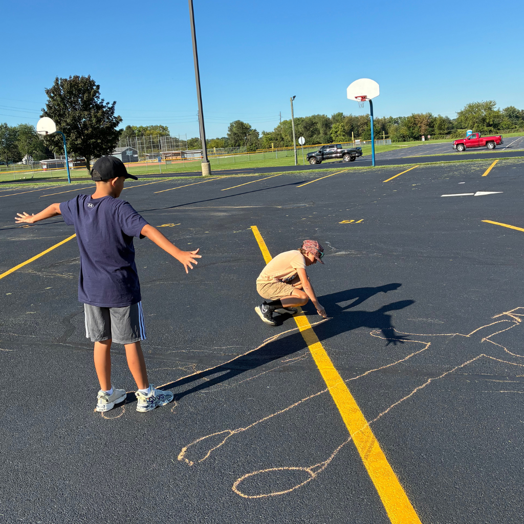 Boy wearing a sports jersey standing on the blacktop while another student crouches to outline his shadow using sidewalk chalk.