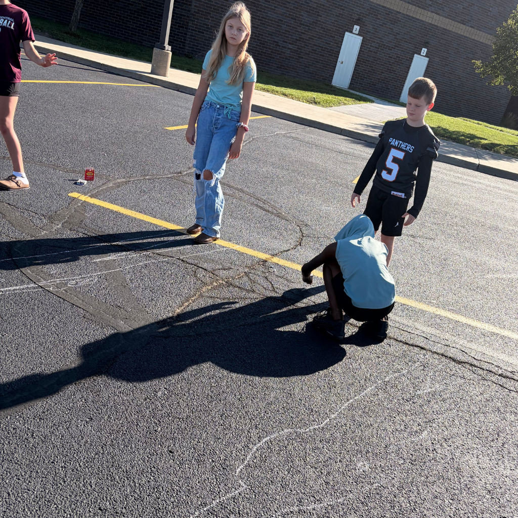 Students participating in an outdoor science activity on the blacktop, using chalk to trace shadow outlines cast by classmates.