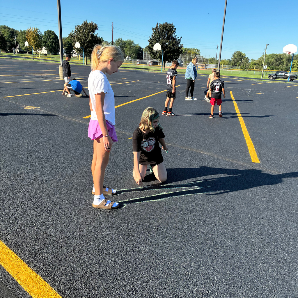 Young girl standing on the school blacktop while a classmate kneels down to trace her shadow with sidewalk chalk on a sunny day.