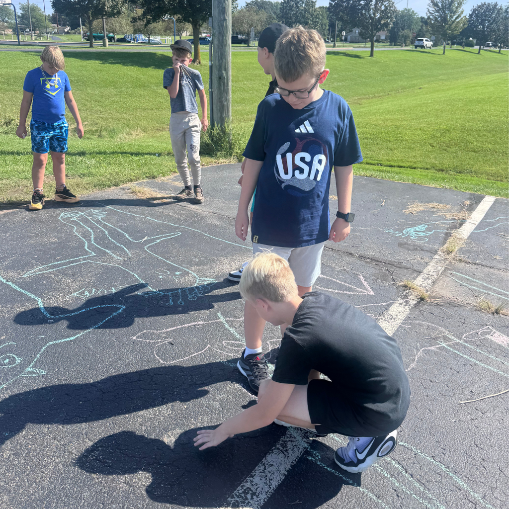 Elementary students gathered on the school blacktop, drawing and tracing shadow figures on the asphalt using colorful sidewalk chalk.