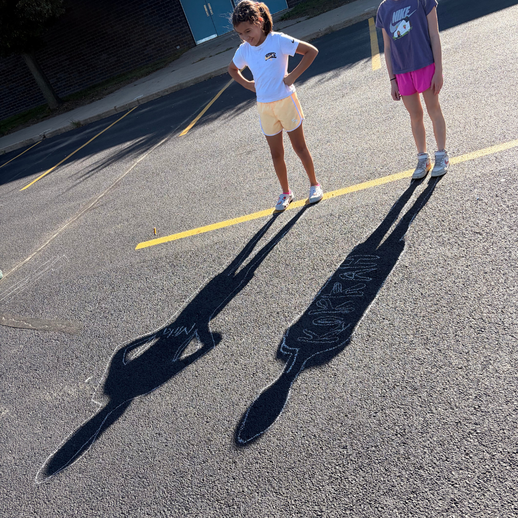 wo young girls standing in a parking lot, inspecting their long shadows on the pavement labeled with chalk names.