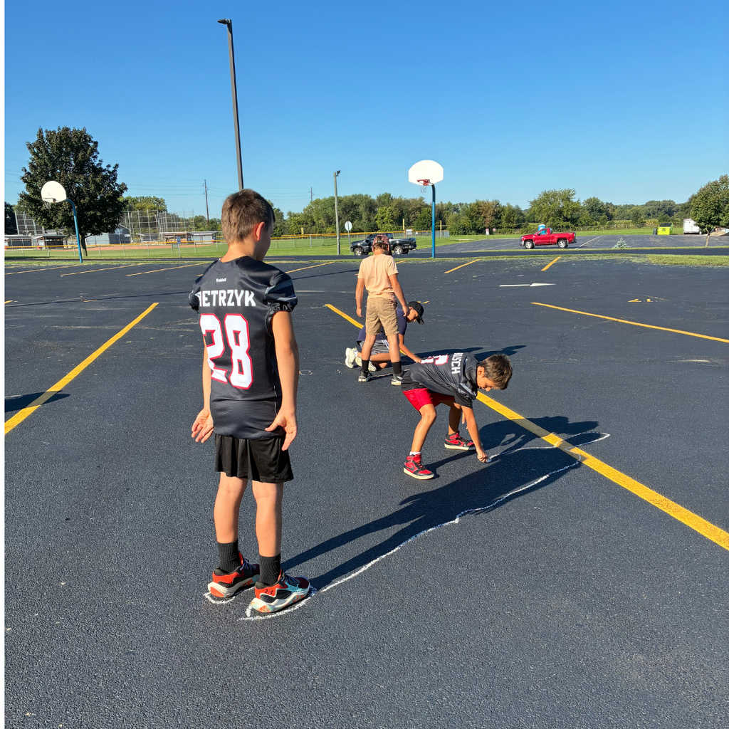 Boy wearing a sports jersey standing on the blacktop while another student crouches to outline his shadow using sidewalk chalk.