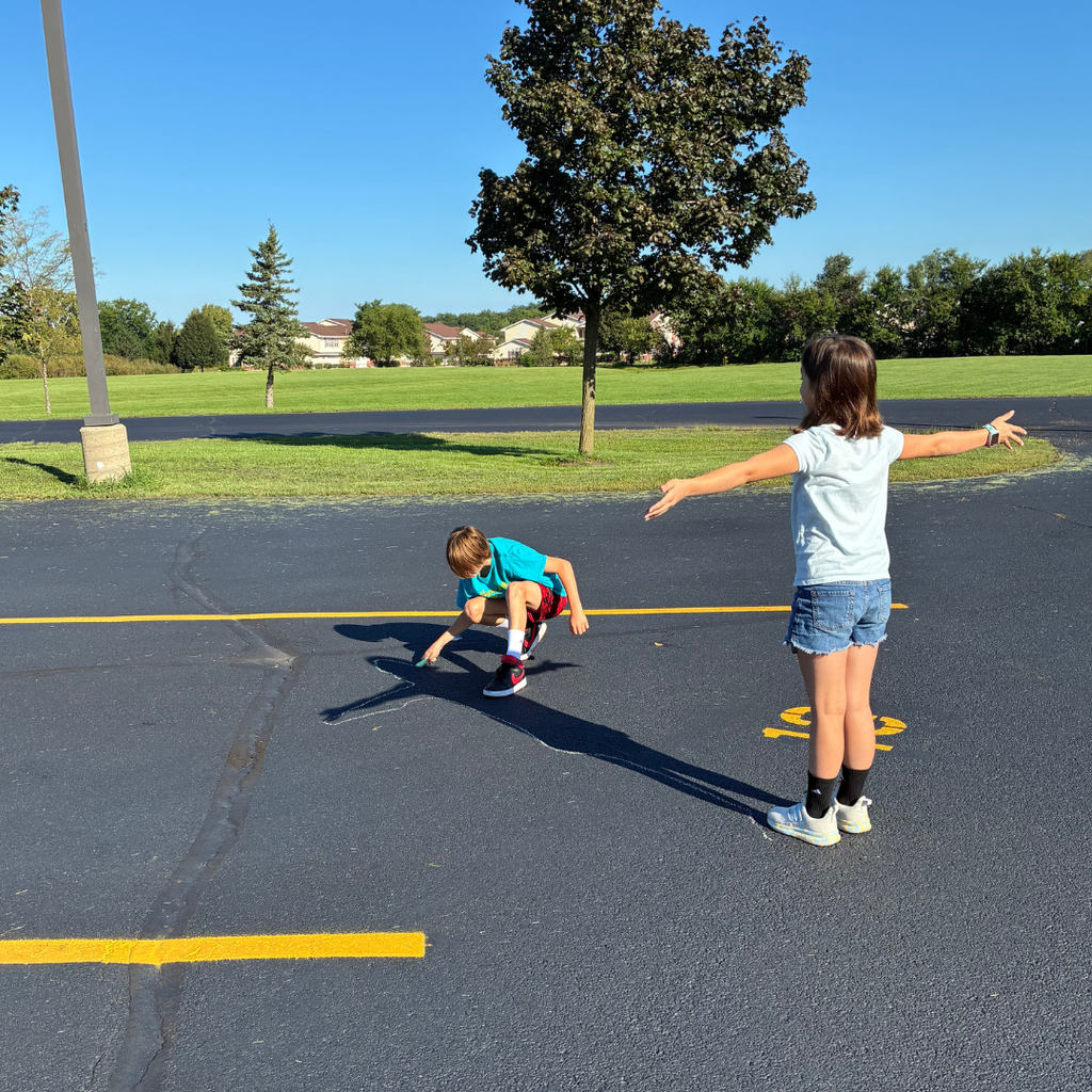Girl posing with arms stretched out on a school blacktop while another student traces her long shadow in sidewalk chalk on a sunny day.