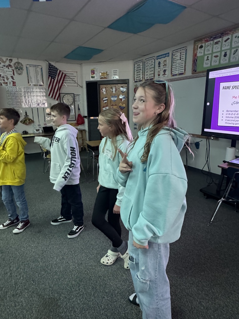 Students smiling and gesturing while participating in a classroom activity near a digital display screen.