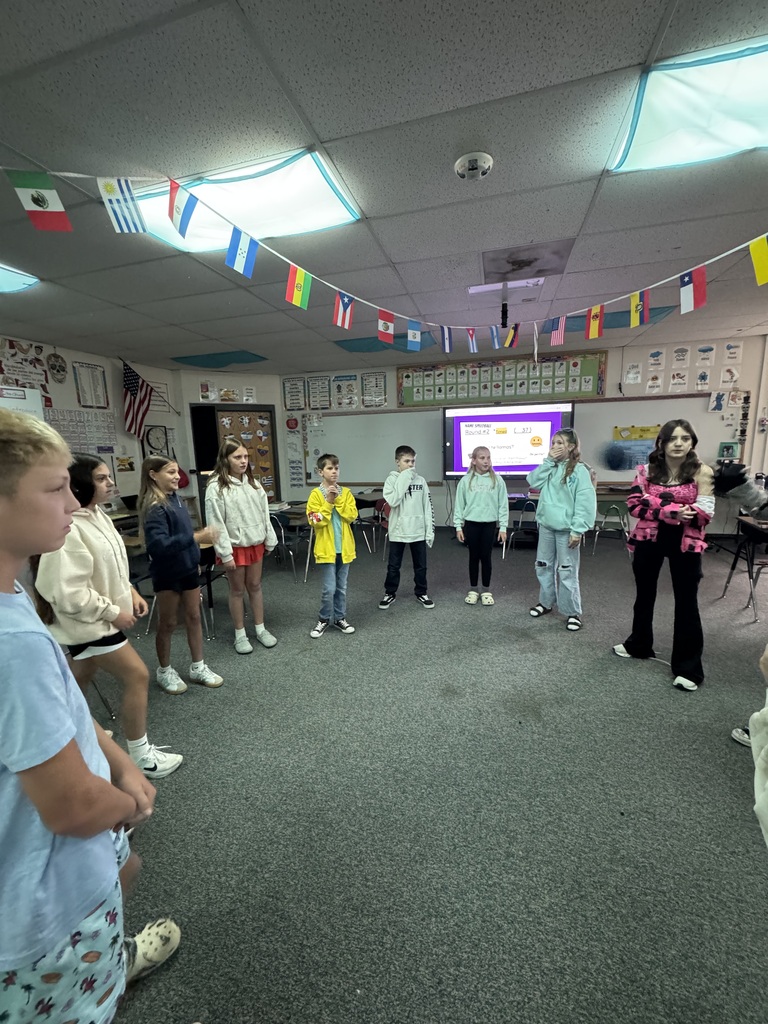 Students playing a ball-tossing icebreaker game inside a Spanish classroom decorated with international flag banners.