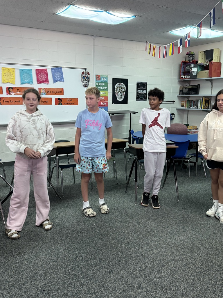 Four middle school students standing side by side in a classroom decorated with Spanish language posters.