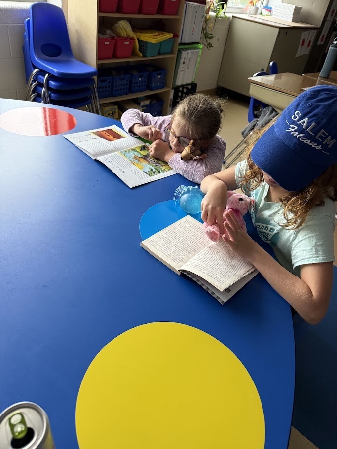 Two young students sitting together at a large blue activity table, silently reading books.