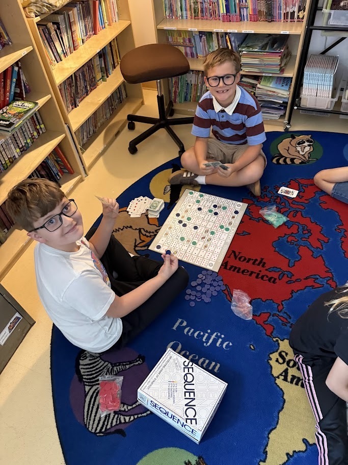 Two young boys smiling while playing the Sequence board game on a world map rug in a classroom library.
