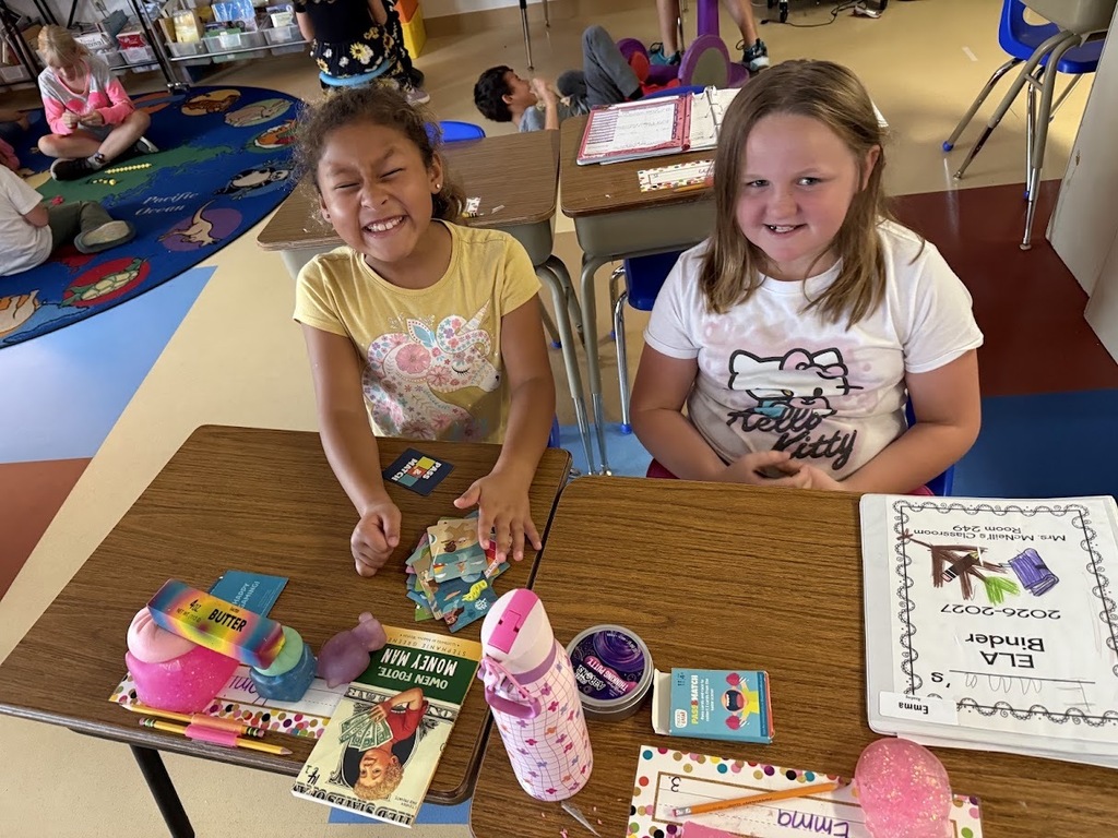 Two young students sitting at desks in a classroom, smiling with activity cards and school supplies spread out.