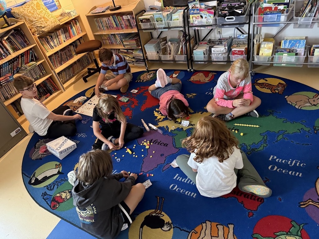 Group of elementary students sitting in a circle on a world map rug, playing board games in the classroom.