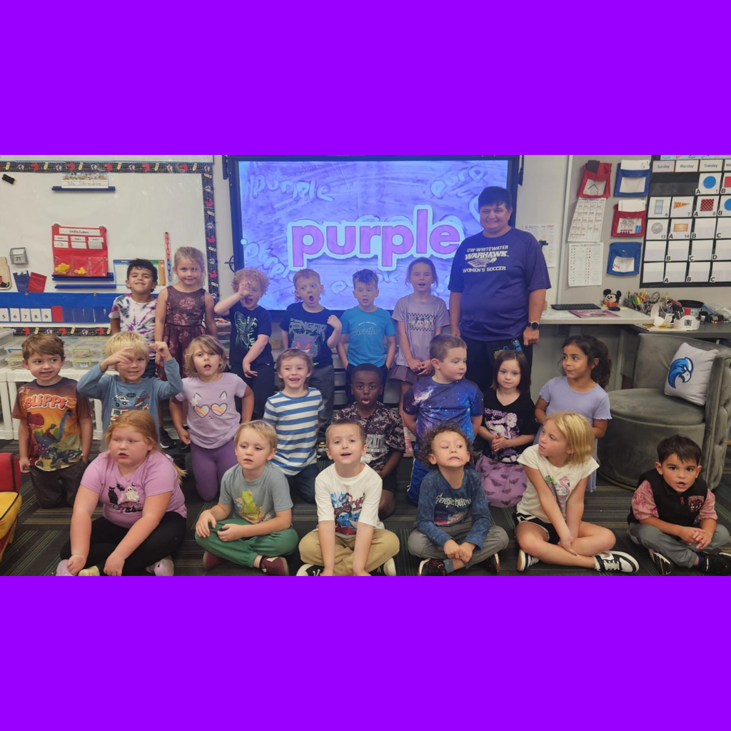 Elementary class wearing purple outfits posing in front of a classroom screen displaying the word "purple."