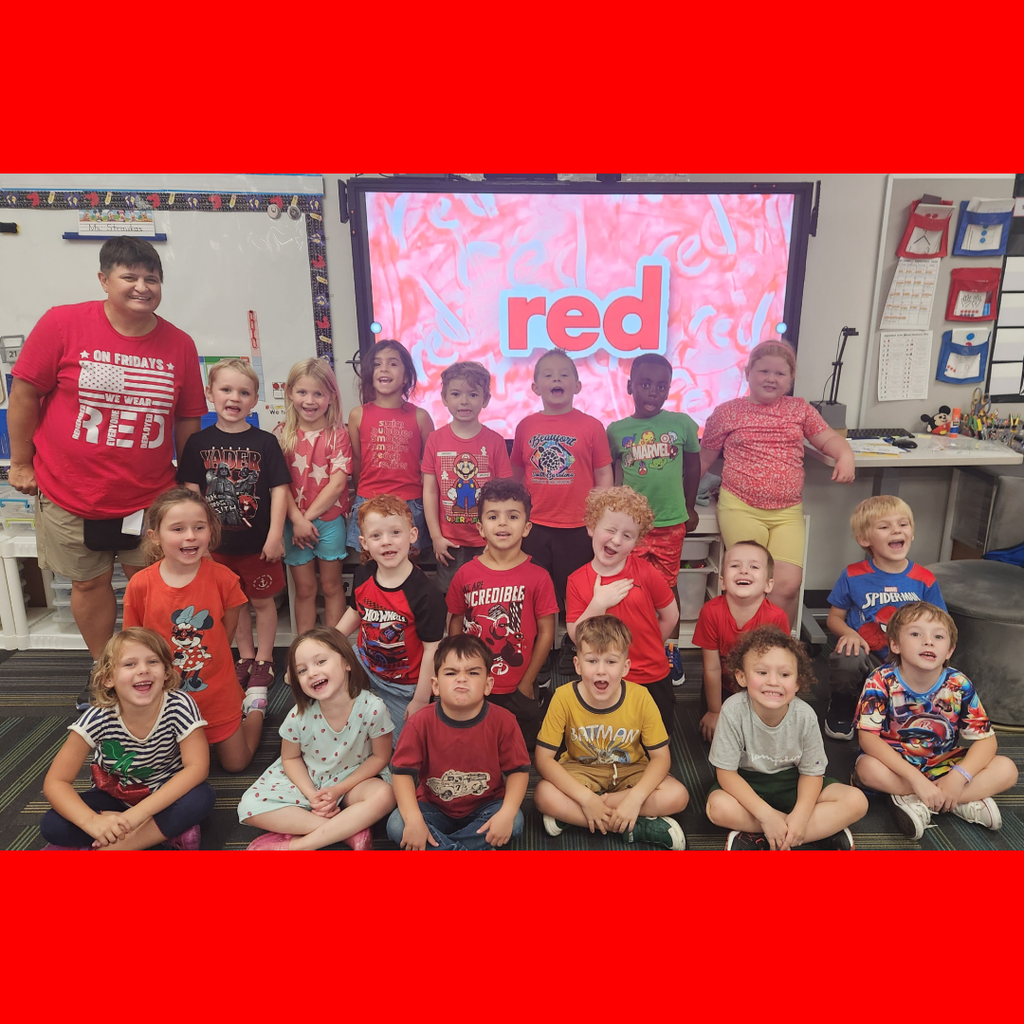 Classroom of elementary students dressed in red clothes posing together in front of a screen reading "red."