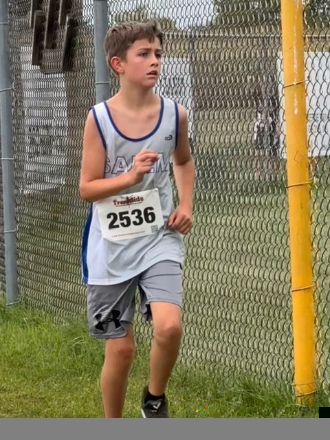 Boy wearing a Salem athletic tank and race bib 2536 running next to a chain-link fence.