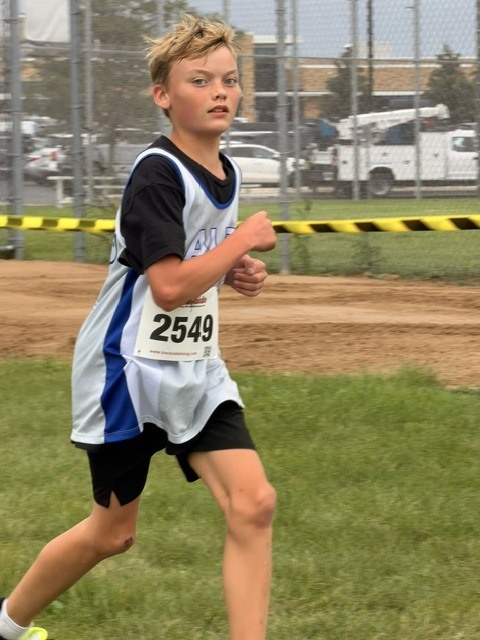 Young boy wearing bib 2549 actively running on a cross country course near a fence.