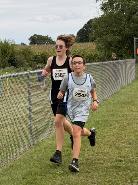 Two middle school runners wearing race bibs competing along a fenced grass path.