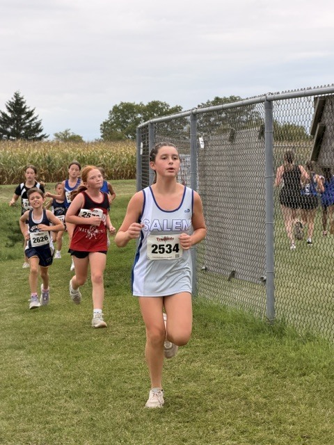 Group of student runners in uniform competing during an outdoor cross country race along a chain-link fence.