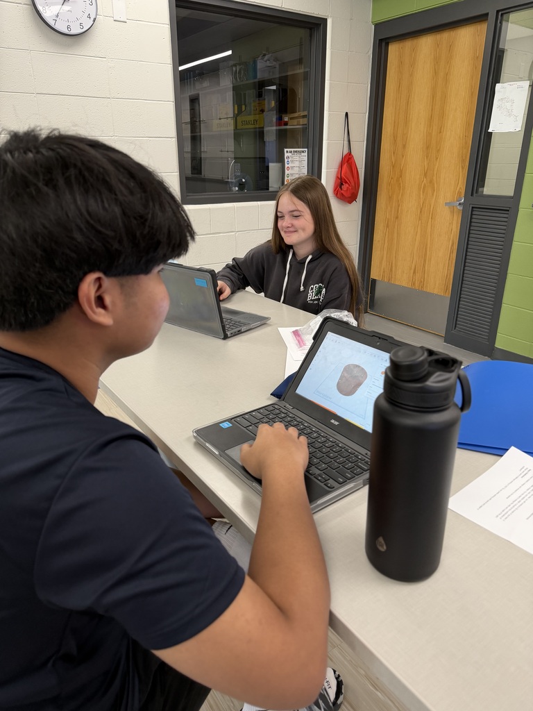 Two students collaborate at a classroom table while working on 3D design software on their laptops.