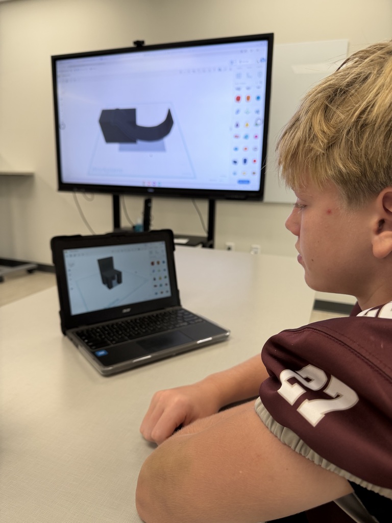 A student wearing a sports jersey works on a 3D modeling design on a laptop in front of a digital display monitor in a classroom.