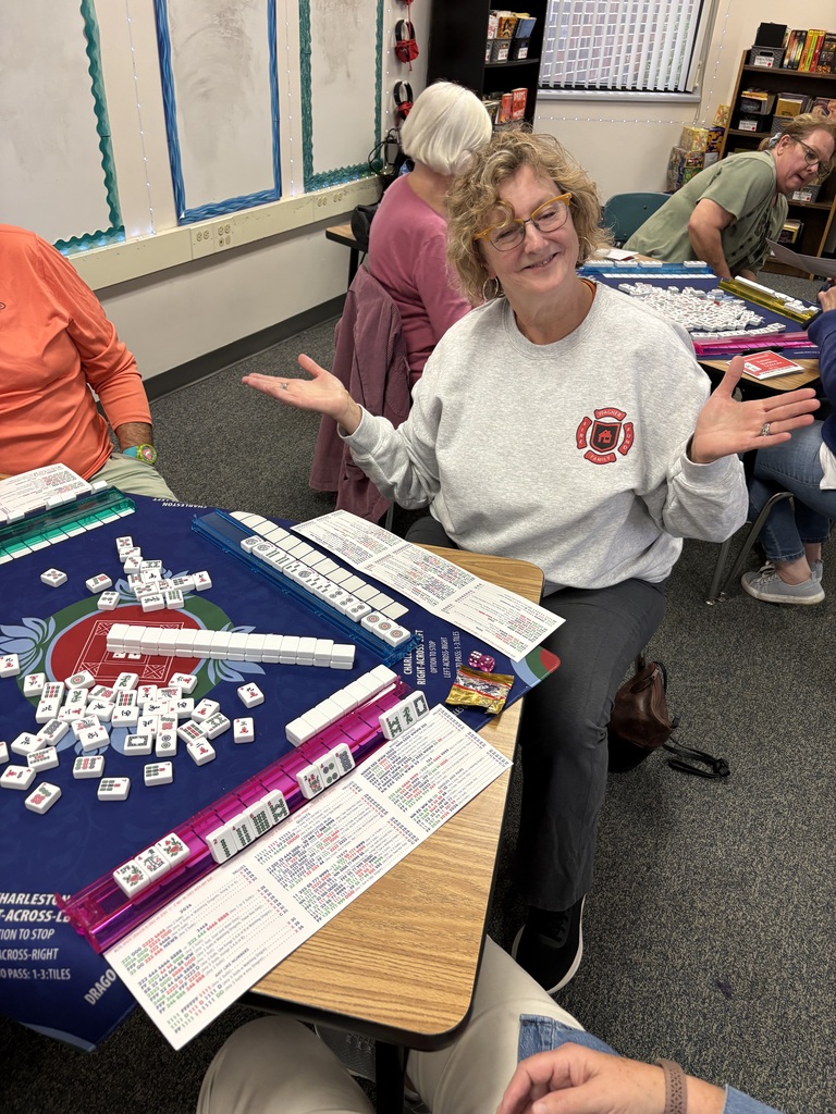 A woman with curly hair shrugging with open hands while seated at a table playing Mahjong