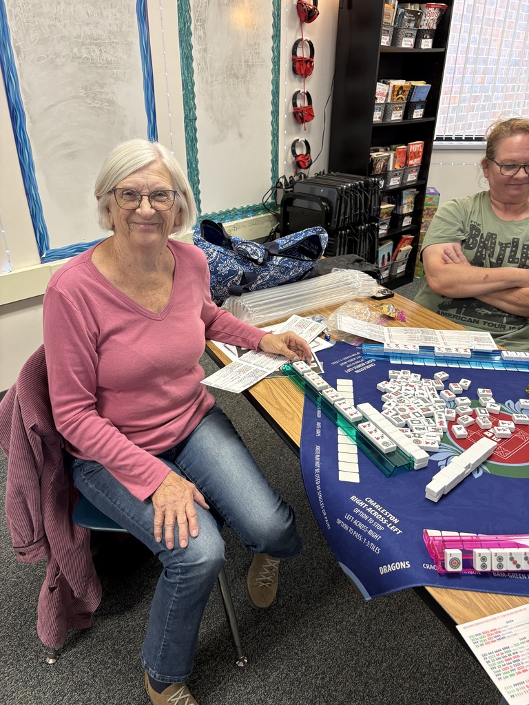 A smiling woman in a pink long-sleeve shirt sitting at a desk with a Mahjong tile game set up in front of her.
