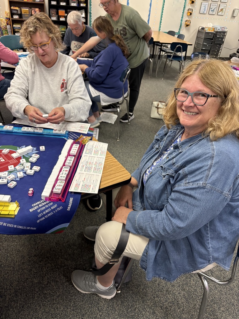 A smiling woman with glasses and a denim jacket seated at a table during a community Mahjong game night.